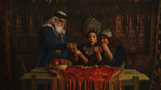 A staged photograph by Reza Daryakenari: an older man with a white beard offers grapes to a woman at a table covered in red embroidered cloth, a second woman looks on, and a bowl of grapes and pomegranates stands on the table.