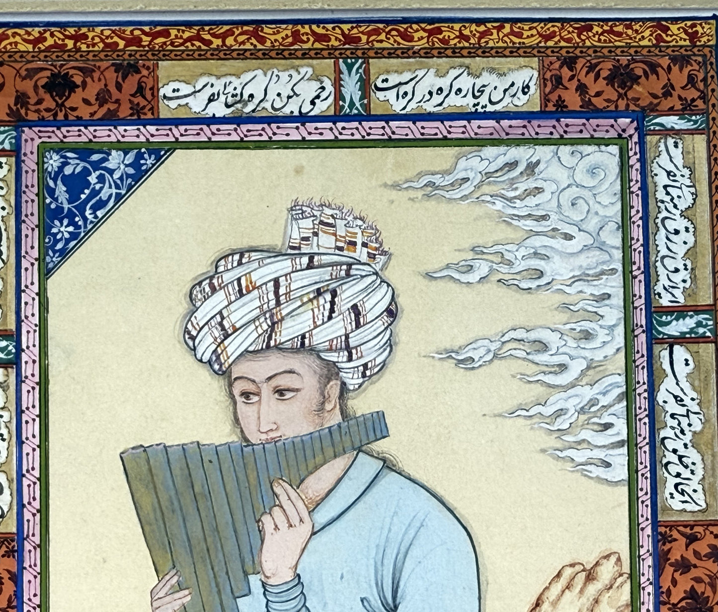 Detail: the musician's face under a striped white turban, lips at grey-blue panpipes, with Chinese-style cloud scrolls drifting past and a blue-and-white floral corner panel - Muraqqa': The Blue Robe, Kamal al-Din Ghotb.