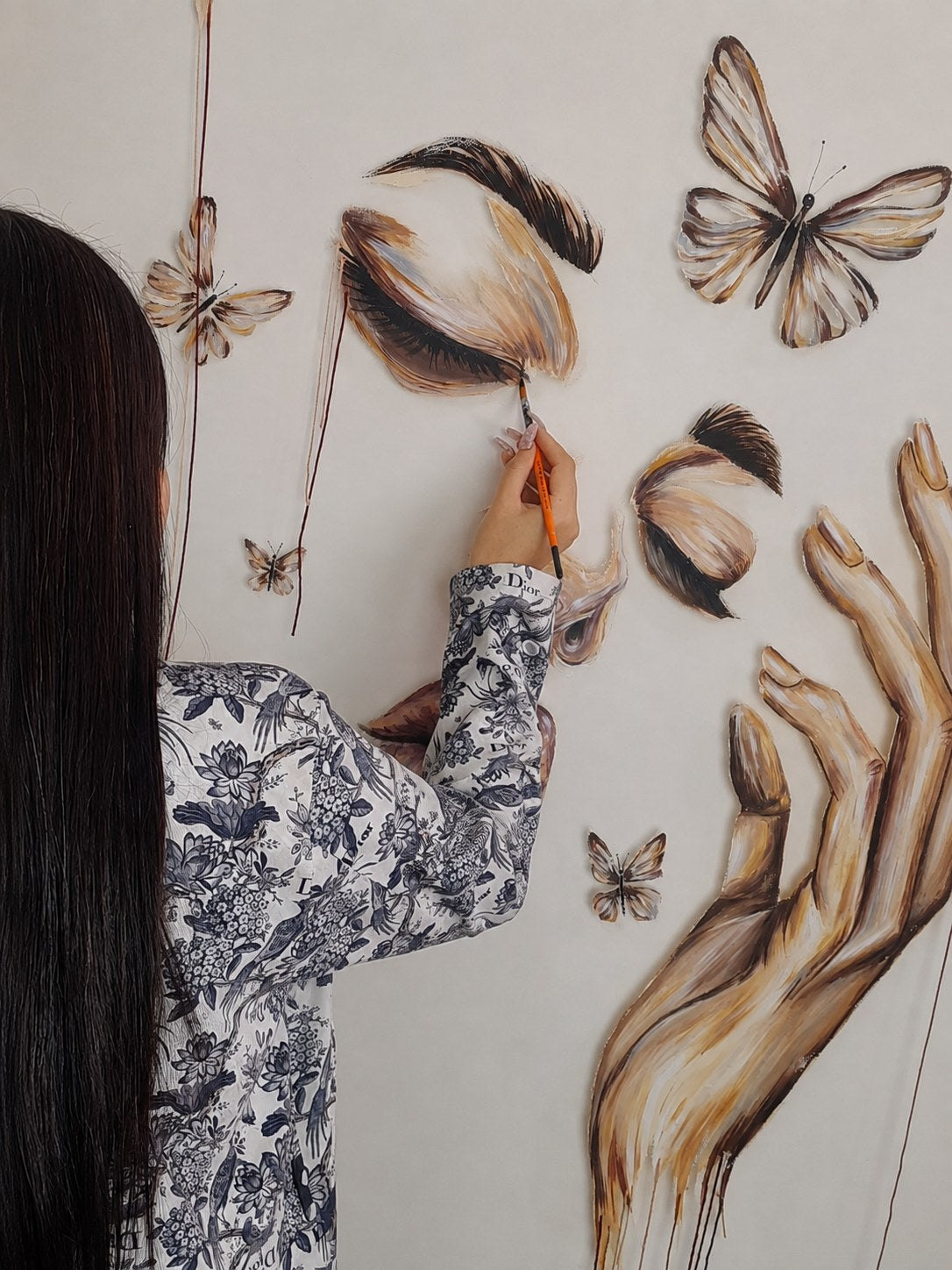 Threads of Becoming by Mahdiye in progress: the artist, in a blue-and-white floral jacket, painting the upper closed eye among butterflies and a reaching hand